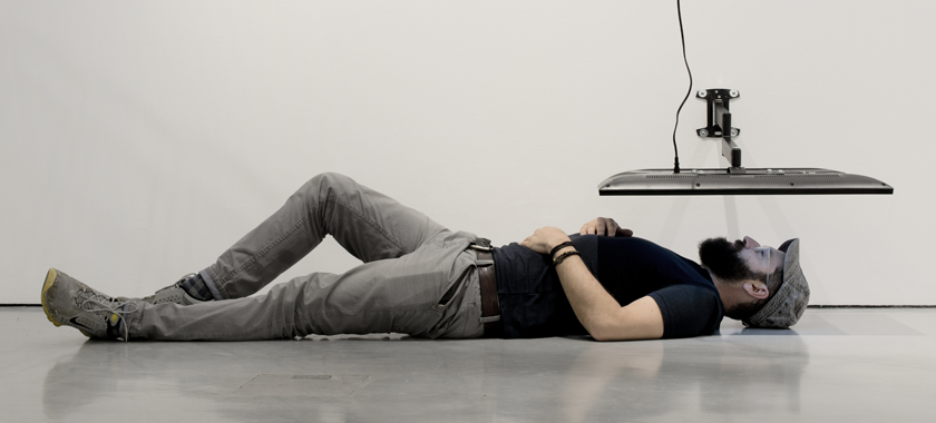 Image Detail: an individual is laying on their back on a smooth concrete floor, their head under a TV monitor that is mounted to the wall, screen facing down towards their face.