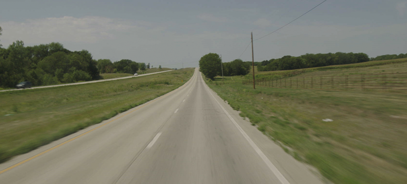 A head-on windshield view from an 18-wheeler of a single-lane country road with a clear blue sky surrounded by green trees and fields