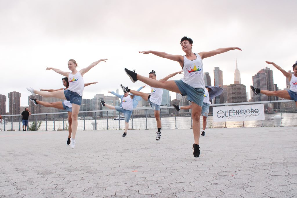 A group of dancers wearing white tank tops and black shorts pose in a dance move, with an audience watching in the background.