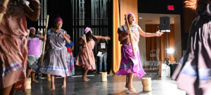 Women singing and dancing on a stage, holding mortar and pestle tools used to mash plantains.