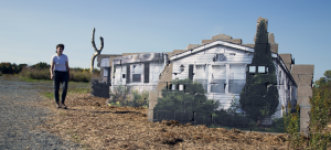 Three cinderblock walls with images of mobile homes pasted to each facade. They are situated in a large pastoral sculpture park with the artist walking next to them wearing a white shirt and black jeans.