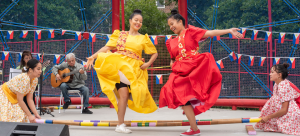 Two dancers in center stage, dressed vibrantly, with musicians in the background and additional performers on either side